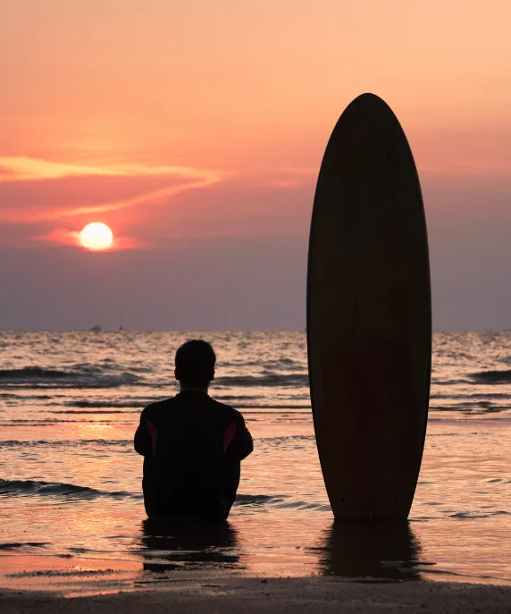 Surfista sentado na beira do mar ao lado da prancha, contra o pôr do sol