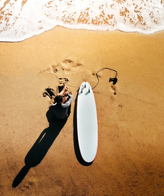 Vista aérea de um surfista sozinho na areia ao lado da prancha, com a espuma da onda chegando